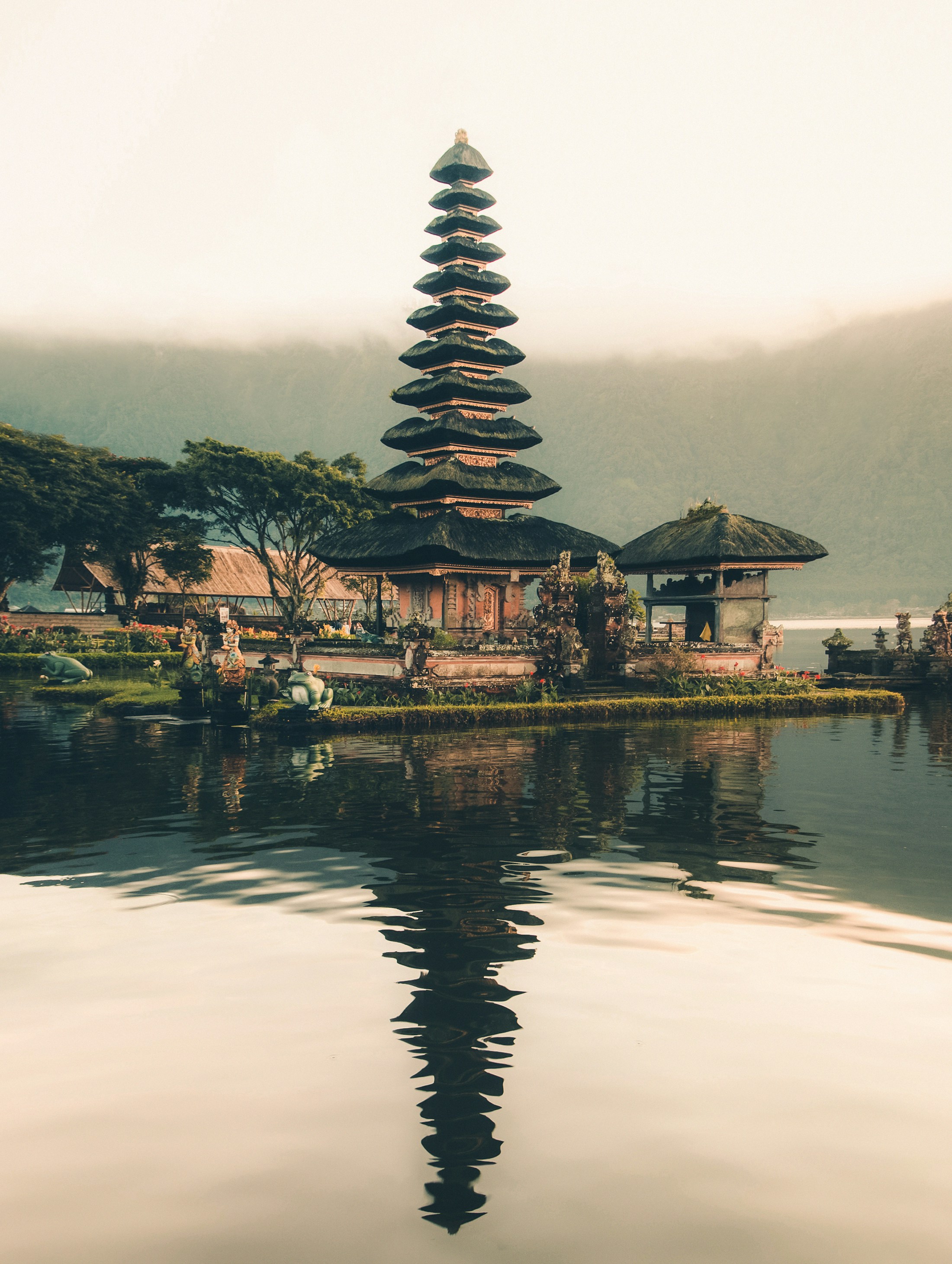 A Balinese lakeside temple reflected in calm water, with mountains beyond